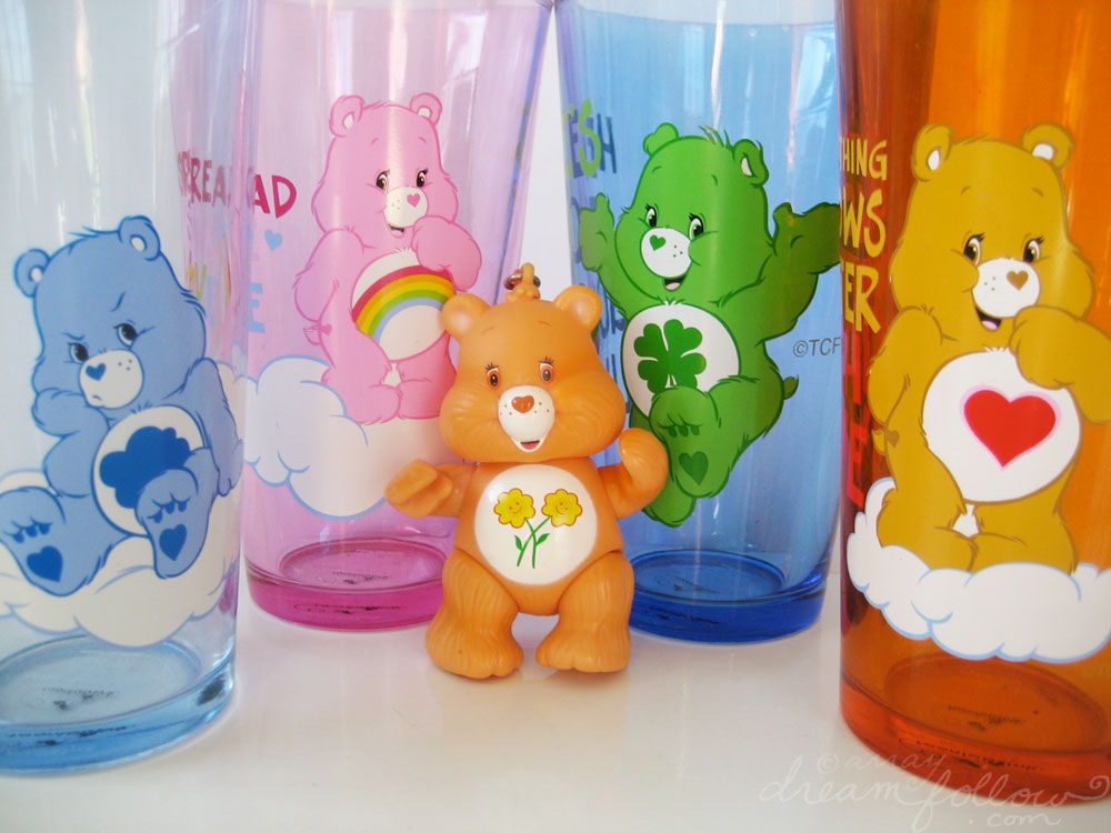 Friend Bear, the light-orange bear, with two smiling sunflowers on her tummy, with four Care Bear tumblers at the back