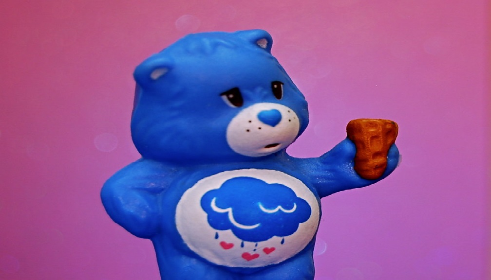 Grumpy Bear, an indigo blue Care Bear, holding an ice cream cone; on his tummy is a raincloud with falling raindrops and hearts.