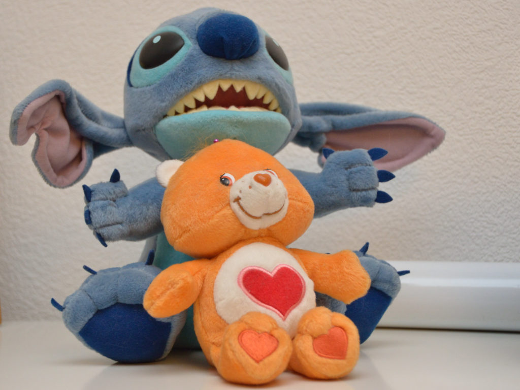 Tenderheart, an orange-brown Care Bear with a red heart with a pink outline on his tummy; on his back is a bigger Stitch stuffed toy