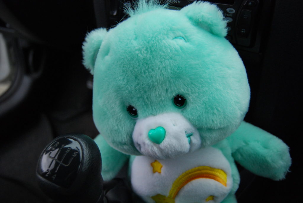 Teal Care Bear named Wish Bear inside a car; on her tummy is a smiling yellow shooting star with a gold comet tail surrounded by three other smaller stars