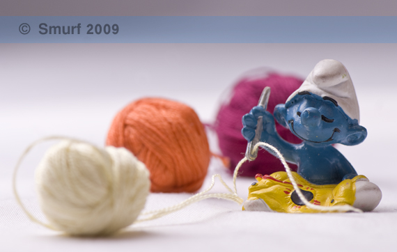 Tailor Smurf sitting down while sewing a dress using three different colors of yarns