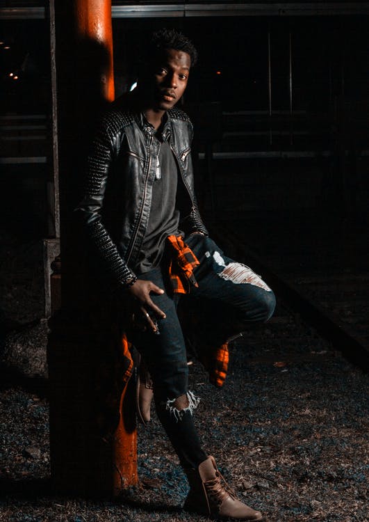 A man wearing a black leather jacket and distressed jeans while reclining on a red  post