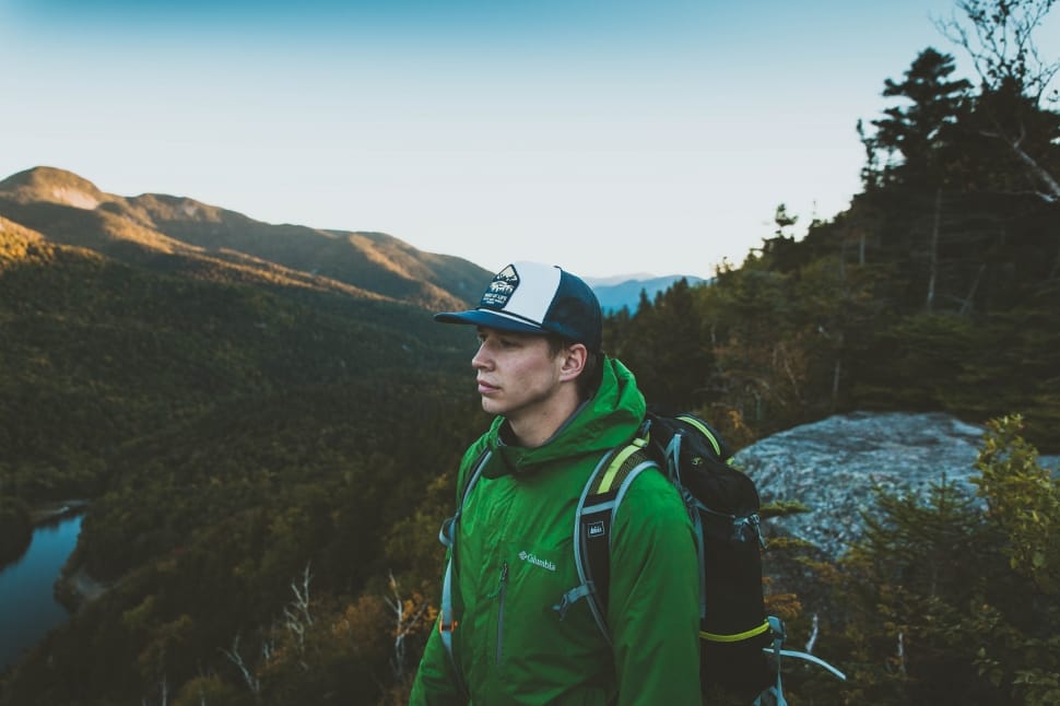 A man at the peak of a mountain wearing a cap and a green windbreaker