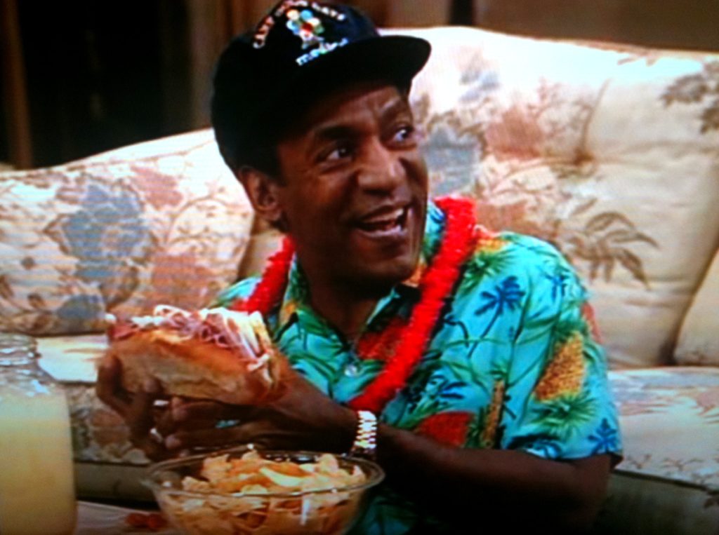 Bill Cosby in Hawaiian polo and black cap while holding paper bills 